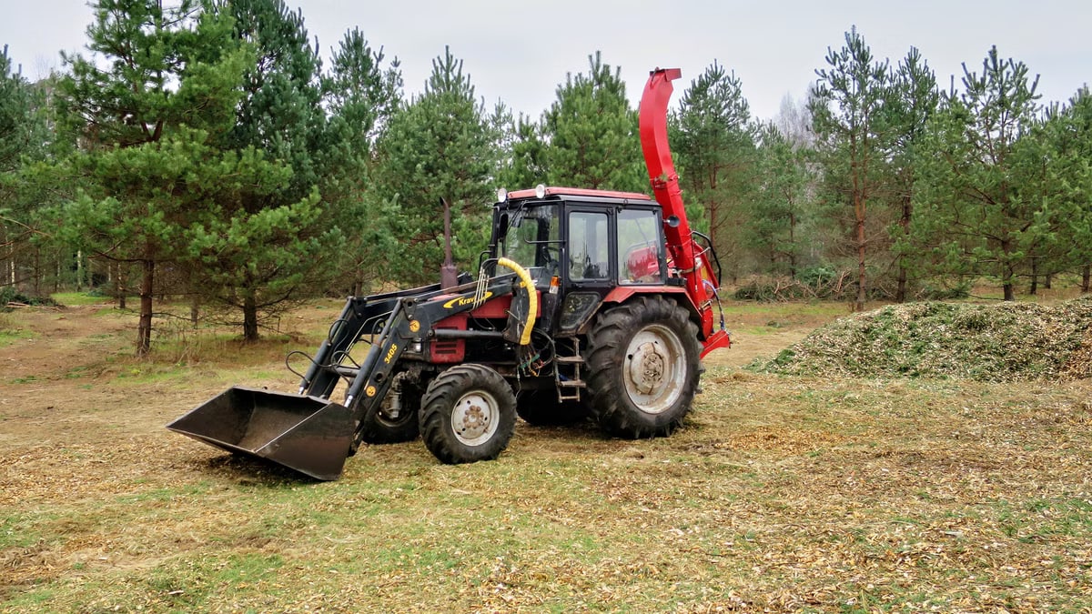 A red tractor equipped for logging and wood chipping, set in a dense forest.